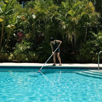 Personne nettoyant une piscine avec une brosse longue portée, eau claire, lumière du soleil